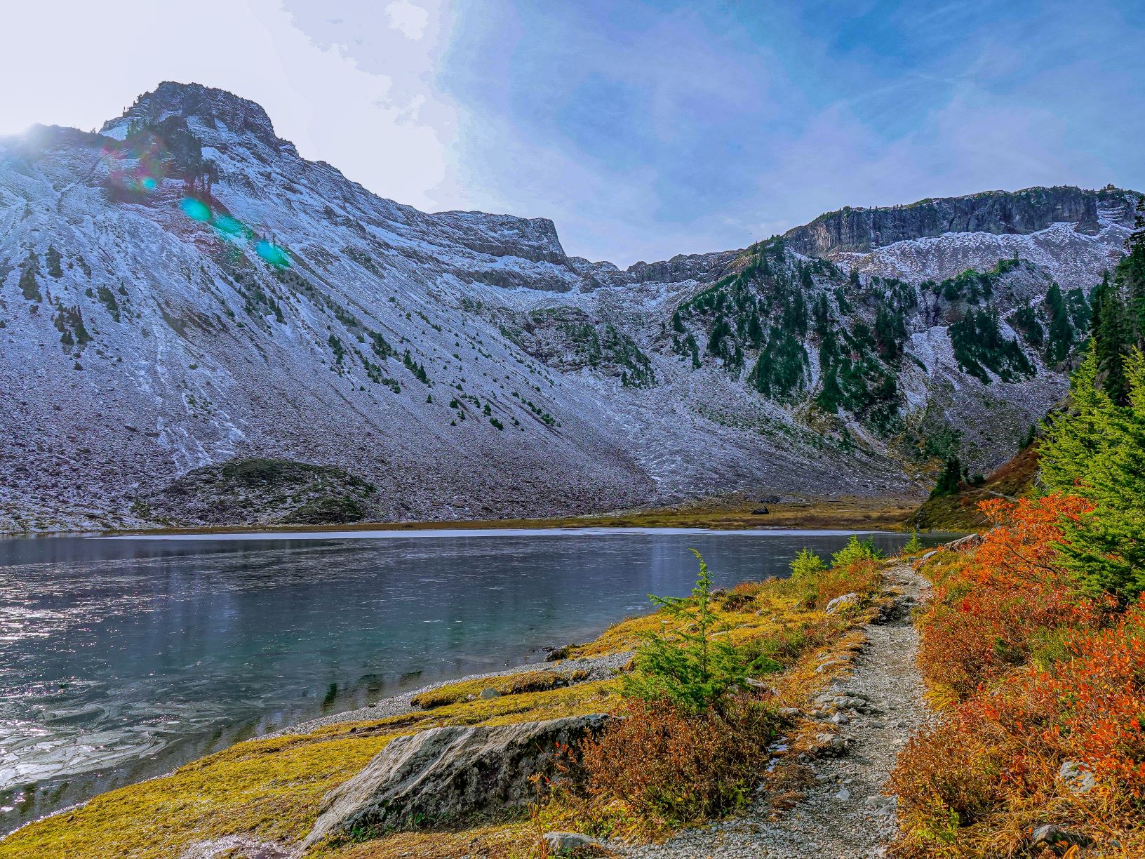 Mountain in background with dusting of snow. Water through the middle of the image. A small dirt pathway and colorful plants in the foreground.